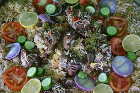 Large platter of yellowish kabsa rice with bone-in meat, tomato, cucumber, onion and lemon slices