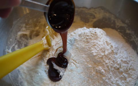 Dark malt syrup poured from a measuring spoon onto flour in a mixing bowl