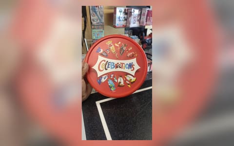 A Celebrations tub held up in a shop, its lid showing Snickers, Twix, Maltesers, Bounty and Galaxy wrappers