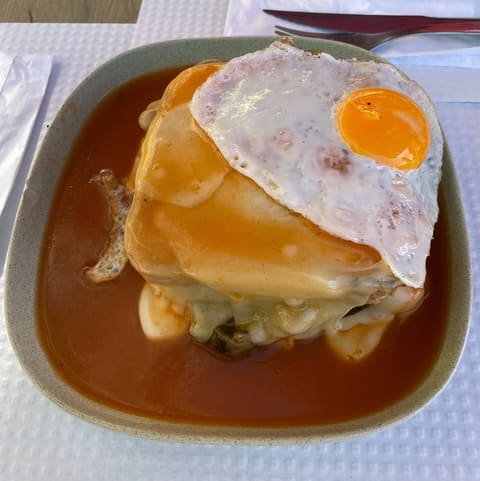 Porto francesinha covered in melted cheese and orange sauce, topped with a fried egg in a square bowl